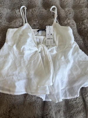 Vit omlottopp med knyt från Gina Tricot - Säljer en luftig vit omlottopp från Gina Tricot i linne med smala axelband och knyt framtill. Toppen har en flowy passform och är perfekt för varma dagar. Supersnygg till både jeans och kjol!
