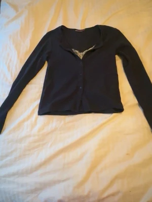 Svart cardigan med spetsdetalj - Snygg svart cardigan med långa ärmar och knäppning framtill. Spetsdetalj vid halsringningen ger en extra touch. Perfekt att slänga över ett linne eller topp för en chill look.  Kofta  ifrån gina 14+ med inbyggt linne, väldigt bra skick 