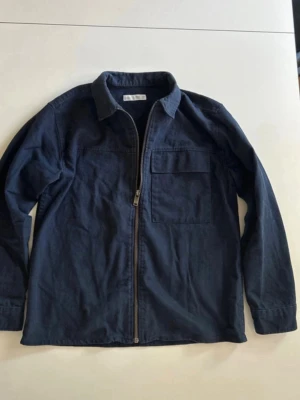 Mörkblå overshirt med dragkedja - Snygg mörkblå overshirt i bomull med dragkedja framtill och en bröstficka. Klassisk krage och långärmad modell som passar perfekt till lager på lager. Enkel och stilren design som funkar till det mesta. Hör av dig om du har några frågor!