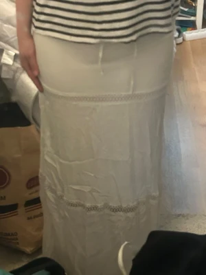 Vit långkjol med spetsdetaljer - Vit långkjol med fina spetsdetaljer och rak passform. Kjolen har en enkel och stilren look med dekorativa spetsband längs kjolen. Perfekt för en somrig och avslappnad stil.