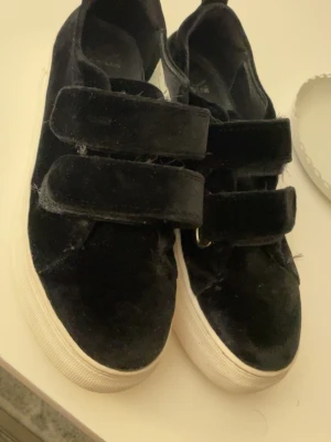 Svarta sneakers - Säljer ett par svarta sneakers med två breda kardborreband och vit tjock sula. Skorna har en mjuk, sammetsliknande yta som ger en cool look. Perfekta för dig som vill ha något enkelt men ändå stilrent till vardags.