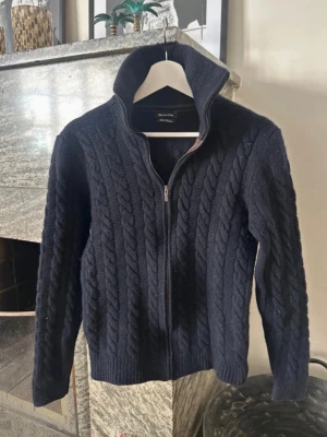 Massimo Dutti Kofta - Mörkblå kabelstickad kofta från Massimo Dutti med dragkedja framtill och hög krage. Koftan har ribbstickade muddar och en klassisk, tidlös design som passar perfekt till lager-på-lager. Materialet ser ut att vara ullblandning för extra värme. Passar 160-170cm
