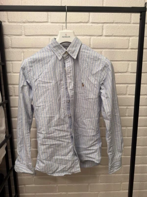 Blårandig skjorta från Polo Ralph Lauren - Säljer en blårandig skjorta från Polo Ralph Lauren med klassisk krage och lång ärm. Skjortan har vit knappstängning och den ikoniska broderade loggan på bröstet. Modellen är custom fit och materialet är mjuk bomull.