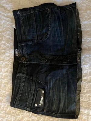 Mörkblå jeanskjol med knappar - Snygg mörkblå jeanskjol från ZERO ONE med rå kant nertill, dekorativa knappar framtill och klassiska fem fickor. Tuff tvättad denim med detaljer som kontrastsömmar och små dragkedjor på bakfickorna. Perfekt för en trendig och avslappnad look.