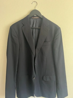 Mörkblå kavaj från Oscar Jacobson - Stilren mörkblå kavaj från Oscar Jacobson med klassisk passform, två knappar framtill och tre fickor. Tillverkad i ett slätt tyg med diskret glans. Perfekt för en dressad look och enkel att matcha med skjorta eller t-shirt.