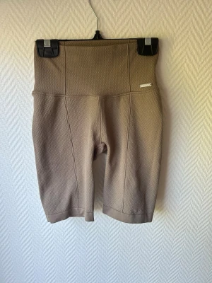 Beige högmidjade cykelshorts - Säljer ett par beige högmidjade cykelshorts i ett strukturerat material. Shortsen har en bred linning och sitter tight för en snygg siluett. Perfekta till sommaren och träning. Inga fickor eller dragkedjor.