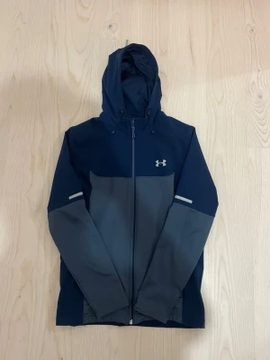 Blå och grå vindjacka Under Armour - Snygg vindjacka från Under Armour i marinblått och mörkgrått. Jackan har huva med dragsko, hel dragkedja framtill och två sidofickor med dragkedja. Reflekterande detaljer på ärmarna och logga på bröstet. Perfekt för blåsiga dagar.