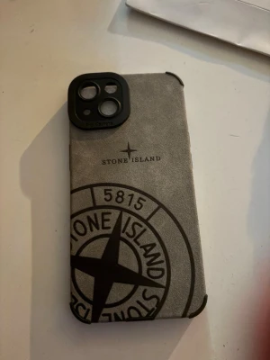 Grått Stone Island mobilskal - Snyggt mobilskal i grått med Stone Island-logga och svart kant. Skalet har en stilren design med tydlig logotyp och passar perfekt för dig som vill ha en unik look på mobilen. Materialet känns tåligt och ger bra skydd mot repor.