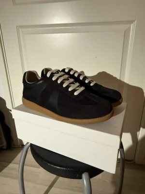 Maison Margiela Svartaoch Bruna 43 - Stilrena svarta sneakers med beige gummisula och vita snören. Skorna har en klassisk låg profil och är tillverkade i en mix av mocka och läder. Perfekta för dig som gillar minimalistisk och tidlös design.