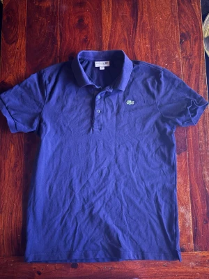 Mörkblå pikétröja från Lacoste - Klassisk mörkblå pikétröja från Lacoste med korta ärmar och den ikoniska gröna krokodilloggan på bröstet. Tröjan har krage och knappar framtill. Perfekt för en clean och stilren look.