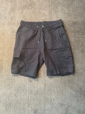 Cargo shorts - BoohooMan  - Säljer ett par stilrena svarta cargo shorts med flera praktiska fickor och justerbar dragsko i midjan. Bekväm passform och en minimalistisk design som passar perfekt till både vardag och streetwear.  • Färg: Svart • Modell: Cargo Shorts • Skick: Nyskick, Inga defekter. • Elastisk midja med dragsko för optimal passform. • Rymliga cargofickor på sidorna samt sidofickor framtill.  📦 Skickas snabbt. Hör gärna av dig om du vill ha fler bilder eller mått.