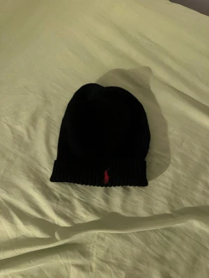 Svart mössa från Polo Ralph Lauren - Svart ribbstickad mössa från Polo Ralph Lauren med klassisk röd logga broderad framtill. Enkel och stilren design som passar perfekt till kalla dagar. Mjuk och bekväm passform.