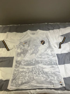 Vit AIK matchtröja med tryck - Snygg vit AIK matchtröja från Nike med detaljerat stadsmotiv i grått på framsidan. AIK:s klubbmärke på bröstet och svarta, gula och vita ränder på ärmsluten. Tillverkad i lätt och ventilerande material, perfekt för träning eller match. En väldigt unik tröja så passa på!
