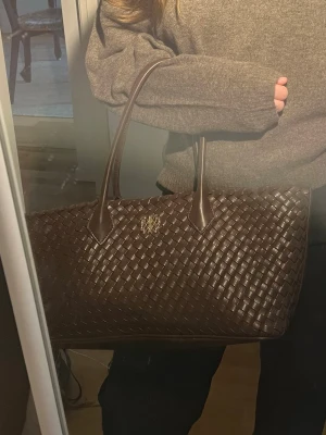 Mörkbrun flätad handväska - Snygg mörkbrun handväska i flätad design med två långa handtag. Väskan har en elegant och stilren look med metallogo framtill. Perfekt storlek för att få plats med allt du behöver. Tillverkad i konstläder med lyxig känsla.