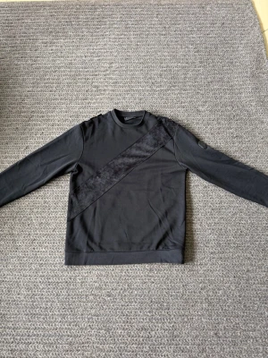 Svart sweatshirt  - Säljer en svart sweatshirt med rund hals och lång ärm. Tröjan har en unik diagonal panel i ett annat material som ger en cool kontrast. Perfekt för dig som gillar stilrena men ändå speciella plagg.