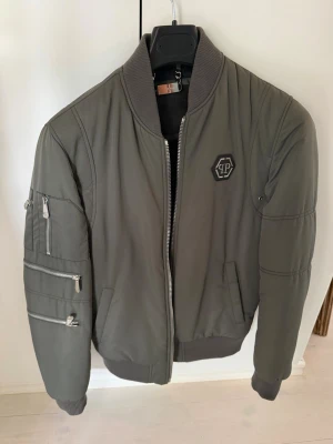 Olivgrön bomberjacka med zip-detaljer - Snygg olivgrön bomberjacka med ribbade muddar och krage. Jackan har flera dragkedjor på ena ärmen och en svart logotyp på bröstet. Klassisk bombermodell med fickor fram och en cool, sportig look. Perfekt för dig som vill ha en stilren jacka med edge.  Vid intresse så går pris att diskuteras då jag aldrig använder denna.
