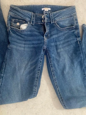 Blå utsvängda jeans från Gina Tricot - Snygga blå jeans från Gina Tricot med utsvängda ben och klassisk femficksmodell. Jeansen har dubbla silverknappar i midjan och vita kontrastsömmar. Tillverkade i mjukt denimtyg som ger en bekväm passform och en tidlös look.