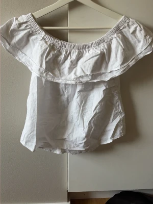 Vit offshoulder topp med volang - Säljer en söt vit offshoulder topp med bred volang runt axlarna. Toppen har en elastisk kant upptill som gör att den sitter snyggt på plats. Perfekt till sommaren och enkel att matcha med olika stilar.