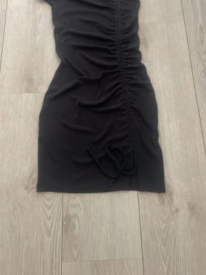 Svart kortklänning med snörning - Snygg svart kortklänning med snörning framtill som ger en cool rynkad effekt. Klänningen har en kroppsnära passform och är perfekt för dig som gillar en stilren look med en twist. Tillverkad i mjukt material som känns skönt mot huden.