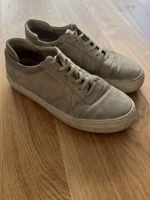 Beige sneakers från Axel Arigato - Snygga beige sneakers från Axel Arigato i mocka med klassisk låg profil och vita sulor. Skorna har tonade skosnören och diskret logga på sidan. Perfekta för en clean och stilren look.