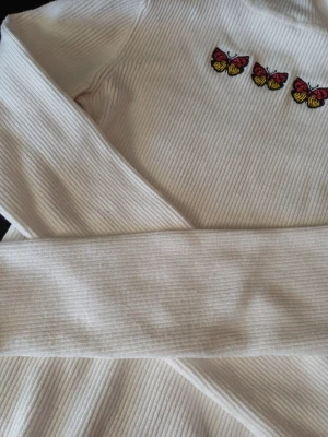 Vit ribbad långärmad topp med fjärilar - Vit ribbad långärmad topp med tre broderade fjärilar i rött, gult och svart framtill. Toppen har en tight passform och är tillverkad i ett stretchigt material som känns bekvämt mot huden. Perfekt för att lyfta en enkel outfit med en söt detalj.
