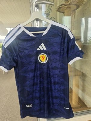  Skottlands hemma fotbollströja Adidas, VM! - Säjer Skottlands VM hemmatröja som är perfekt som minne nu när VM är över. Dessutomdå Skottland gjorde sitt första VM på 28 år! Är I Extremt bra kvalité och jätte snygg. Tröjan har vita detaljer på axlar och ärmslut samt ett diskret geometriskt mönster. Perfekt för dig som vill stötta Skottland på match eller träning.             OBS bara 1 i lager!