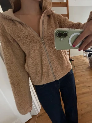 Beige teddyjacka med huva - Mysig beige teddyjacka med dragkedja framtill och huva. Jackan har en avslappnad passform och är gjord i fluffigt, mjukt material som ger en skön och trendig look. Perfekt för kyliga dagar när du vill vara både varm och stylish.