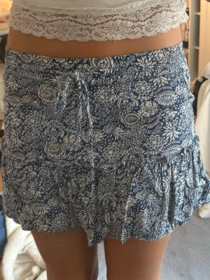 Blåvit kjol med inbyggda shorts - Söt blåvit kjol med blommigt mönster och volangkant. Kjolen har knytband i midjan och är gjord i ett mjukt bomullstyg som känns luftigt och bekvämt. Perfekt för dig som vill ha kjolkänsla men ändå shorts under.