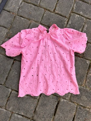 Rosa broderad blus med kort ärm - Söt rosa blus med broderade hål och vågig kant. Blusen har puffiga korta ärmar och liten ståkrage med knapp i nacken. Perfekt för dig som gillar romantisk och feminin stil. Materialet känns som bomull och är lätt och luftigt.