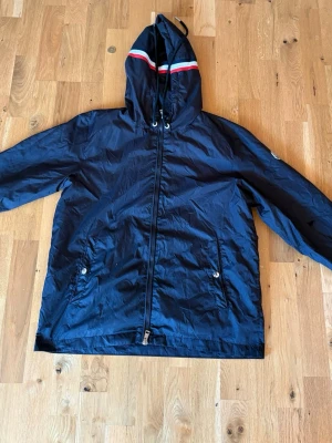 Moncler windbreaker - Snygg marinblå vindjacka från Moncler med huva och dragkedja framtill. Jackan har två fickor med knappar och en ikonisk Moncler-logga på ärmen. Huvan har en röd och vit rand som detalj. Tillverkad i lätt och tåligt material, perfekt för blåsiga dagar. Lite sönder kedja men går att knäppa upp hela vägen