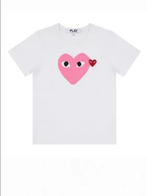 Vit t-shirt med rosa hjärta - Comme de garcon Vit t-shirt med ett stort rosa hjärta med ögon och ett litet rött hjärta på bröstet. Klassisk rund hals och korta ärmar. Perfekt för dig som gillar lekfulla och trendiga prints.