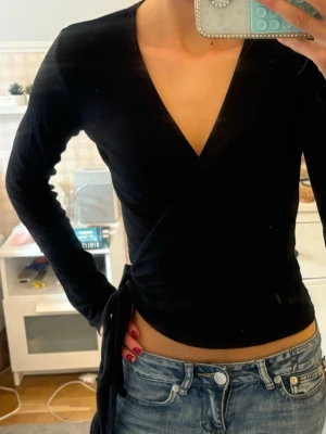 Svart topp med knytband - Snygg svart topp med långa ärmar och knytband i midjan. Toppen har en djup v-ringning och sitter tight mot kroppen, perfekt till jeans eller kjol. Materialet känns mjukt och följsamt.