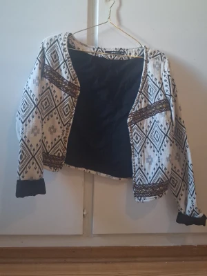 Mönstrad cardiganjacka Gina Tricot - Säljer en snygg cardiganjacka från Gina Tricot i storlek 34. Jackan är vit med svart och brunt etnoinspirerat mönster, har långa ärmar och svarta muddar. Materialet är vävt tyg med dekorativa detaljer och öppen front. Perfekt för dig som gillar unik stil!