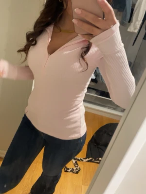 Ljusrosa ribbad långärmad topp - Snygg ljusrosa ribbad topp med lång ärm och v-ringning. Toppen har en slim passform och är tillverkad i ett stretchigt material som sitter skönt på kroppen. Perfekt att styla med jeans eller kjol för en trendig look.