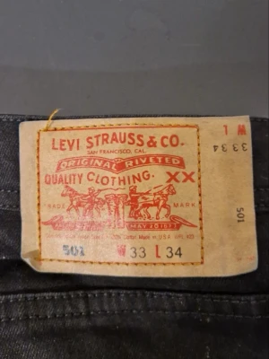 Svarta Levi's 501 jeans - Klassiska svarta Levi's 501 jeans med raka ben och femficksmodell. Jeansen har en ikonisk läderpatch bak i midjan och röd Levi's-tagg på bakfickan. Tillverkade i slitstarkt denimtyg, perfekta för en avslappnad och tidlös stil.