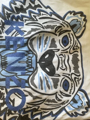 Vit Kenzo t-shirt med tigertryck - Säljer en vit t-shirt från Kenzo med det ikoniska tigertrycket i svart, blått och ljusblått på framsidan. T-shirten har klassisk passform och är tillverkad i mjuk bomull. 