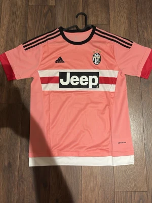 Juventus rosa matchtröja Adidas - Juventus rosa matchtröja från Adidas med svarta detaljer och tre svarta ränder på axlarna. Jeep-logga på bröstet, klubbmärke och Adidas-logga. Tröjan har vita och röda ränder över bröstet samt vit nederkant. Tillverkad i lätt funktionsmaterial.