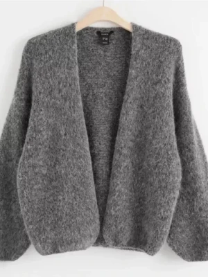 Grå stickad Lindex kofta i mjukt material - Mysig grå stickad kofta från Lindex  med öppen front och lång ärm. Koftan är fluffig och har en avslappnad passform, perfekt att slänga på sig när det är kyligt. Tillverkad i ett mjukt och värmande material som känns skönt mot huden.