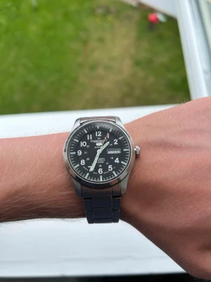 Seiko 5 Sports Automatisk Herr Klocka 100M - Stilren Seiko herrklocka med svart urtavla och tydliga vita siffror. Klockan har en robust länk i silverfärgat stål och dag- och datumvisning. Perfekt accessoar för dig som gillar klassisk och sportig stil.
