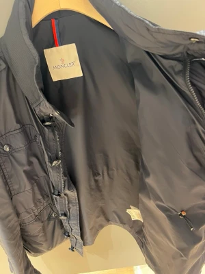 Mörkblå vindjacka från Moncler - Snygg mörkblå vindjacka från Moncler med dragkedja och knappar framtill. Jackan har två bröstfickor med lock, sidofickor och Moncler-logga på ärmen. Tillverkad i lätt, glansigt material som passar perfekt till streetwear och casual outfits.