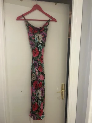 Lång blommig klänning med tunna band - Säljer en lång klänning med tunna axelband och färgstarkt blommönster i rött, lila, turkos, beige och grönt på svart botten. Klänningen har en smal passform och mjukt fallande tyg, perfekt för sommarens alla tillfällen.