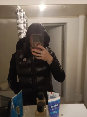 Svart dunväst från Moncler - Snygg svart dunväst från Moncler med glansig finish och klassisk Moncler-logga på bröstet. Västen har en quiltad design och dragkedja framtill. Perfekt för lager-på-lager och ger en cool streetwear-look.