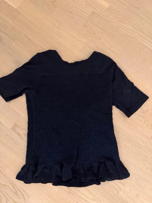 Mörkblå topp med volangkant - Säljer en mörkblå topp med korta ärmar och volangkant nedtill. Toppen har en smockad struktur på framsidan och är tillverkad i ett mjukt, lätt material. Perfekt för dig som gillar en enkel men ändå söt stil.