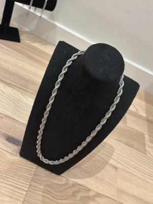 Silver cordell halsband 7mm - Säljer ett stilrent halsband i silverfärg med tvinnad kedja som ger en chunky och modern look. Halsbandet har ett robust lås och passar perfekt för dig som vill ha ett statement-smycke. Materialet ser ut att vara metall och designen är klassisk men trendig.