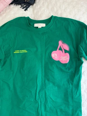 Grön t-shirt med rosa körsbär - Grön t-shirt med stort rosa körsbärstryck och texten 'VIBES ALIGNED, ENERGY MATCHED.' i gult både fram och bak. T-shirten har rund hals och korta ärmar. Perfekt för dig som gillar färgstarka och lekfulla plagg.