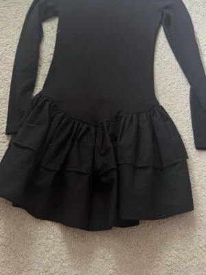 Svart långarmad volangklänning från Zara  - Supersnygg svart klänning med lång ärm och volangkjol i två lager. Klänningen har en tight passform upptill och en kort kjol med volanger som ger en cool och trendig look. Perfekt för dig som gillar stilrena plagg med en twist.