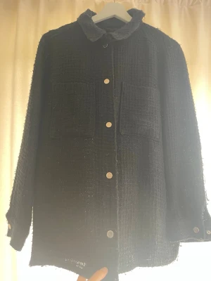 Svart overshirt med fickor - Stilren svart overshirt med grov textur och två stora bröstfickor. Skjortan har tryckknappar framtill och vid ärmsluten samt en klassisk krage.  Mindre i storlek mer i M