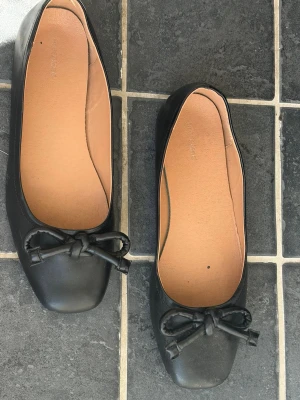 Svarta ballerinaskor med rosett - Stilrena svarta ballerinaskor med fyrkantig tå och dekorativ rosett framtill. Skorna har en slät yta i konstläder och beige innersula för extra komfort. Perfekta för dig som gillar klassisk och enkel stil.