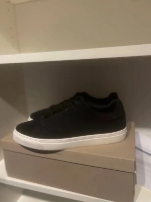 Svarta låga sneakers med vit sula - Stilrena svarta sneakers med lågt skaft och vit sula. Skorna har en klassisk design med svarta snören och ovandel i textil eller mockaliknande material. Passar perfekt till jeans eller chinos för en clean look.