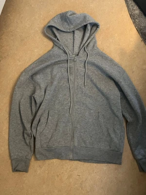 Grå zip hoodie med huva - Klassisk grå hoodie med dragkedja framtill och två fickor. Tröjan har en stor huva med dragsko och ribbade muddar vid ärmar och nederkant. Perfekt för en avslappnad och chill stil. Materialet känns mjukt och bekvämt, troligen bomullsblandning.